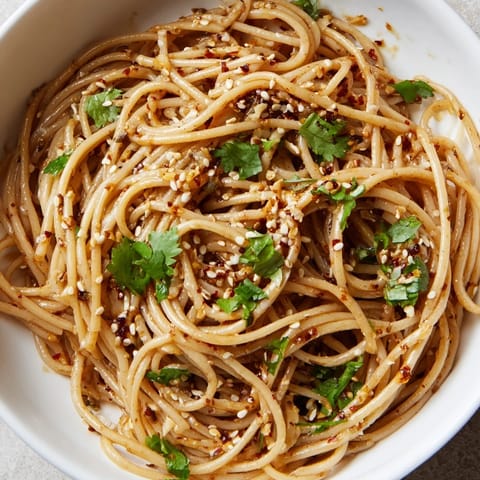 A top-down view of a vibrant Spicy Sesame Noodle Salad featuring chilled wheat noodles tossed in a glossy sesame-chili dressing, topped with julienned cucumber, fresh cilantro, and crunchy roasted peanuts.