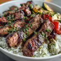 A hearty grilled steak bowl with tender slices of beef, fluffy rice, and colorful roasted vegetables, topped with vibrant chimichurri sauce.
